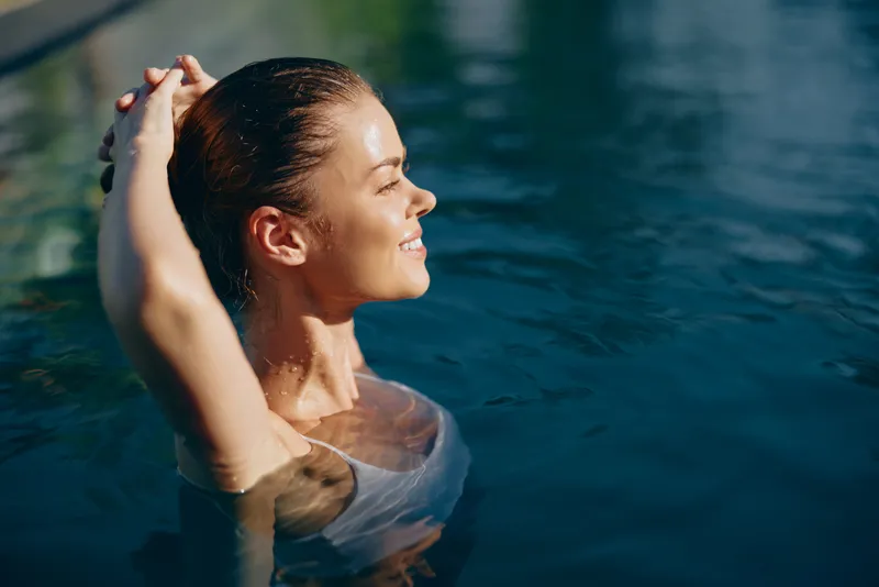 Young woman with wet hair relaxing in outdoor pool water with eyes closed and hands behind head. Concept of lifestyle, wellness, and summer leisure in natural light.