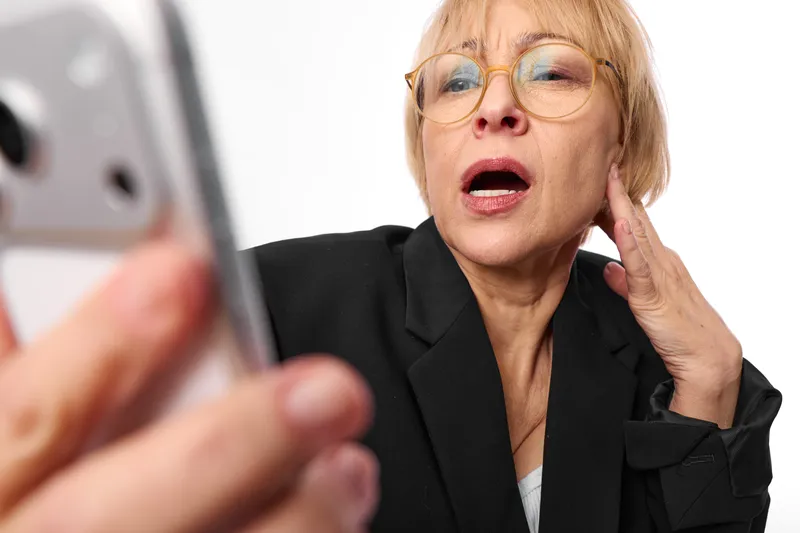 Elderly woman business suit glasses holds phone, smartphone call, surprised closeup portrait, communication with blurred device foreground office assistant
