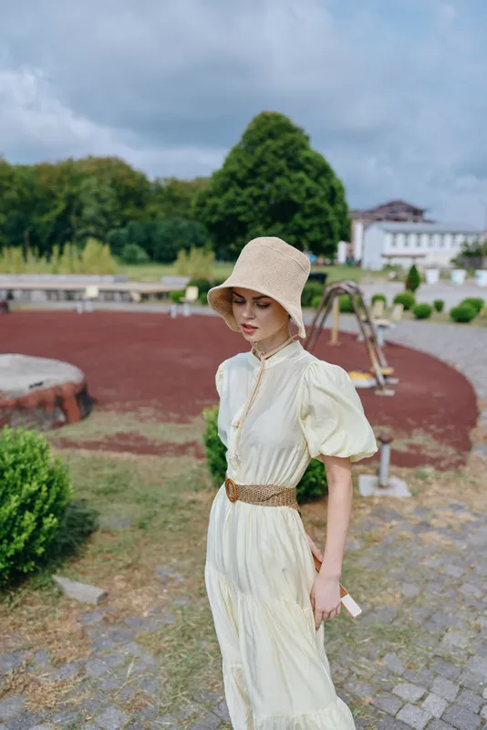 Elegant woman in a cream dress and straw hat stands outdoors in a manicured garden, showcasing vintage fashion and serene summertime ambience with soft sunlight and tranquil surroundings.