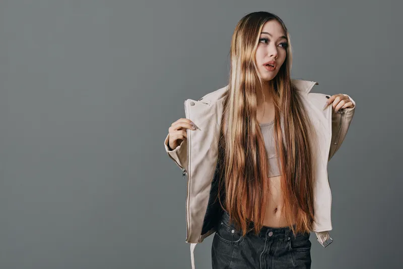 Young woman with long hair wearing light jacket and dark pants posing in studio on gray background. Fashion, casual style and confident look with natural makeup and loose hair.