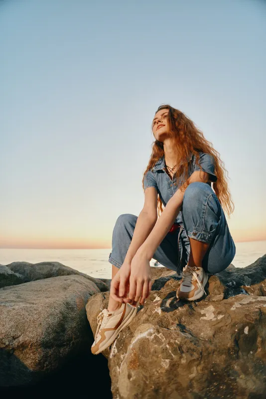 young woman sitting on rocks by the sea during sunset wearing denim casual clothes and sneakers with relaxed and happy mood