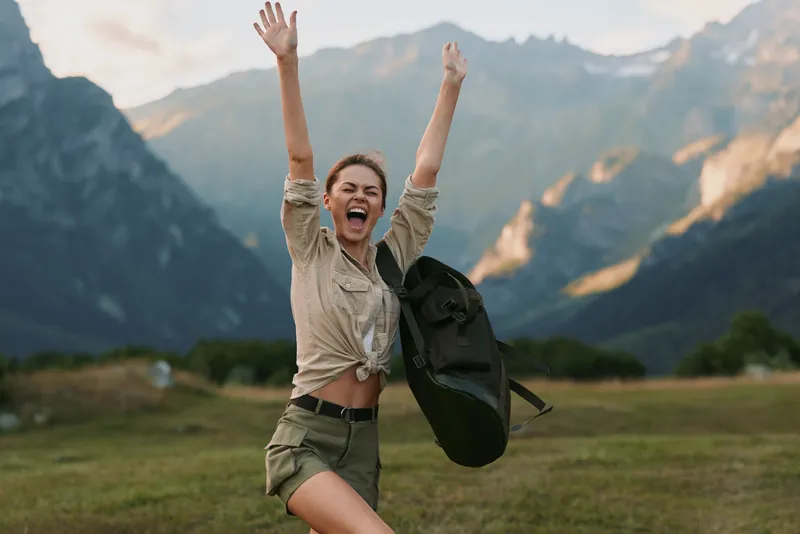 Young woman with backpack raises arms in triumph in a grassy valley beneath towering mountains, conveying adventure and outdoor freedom.