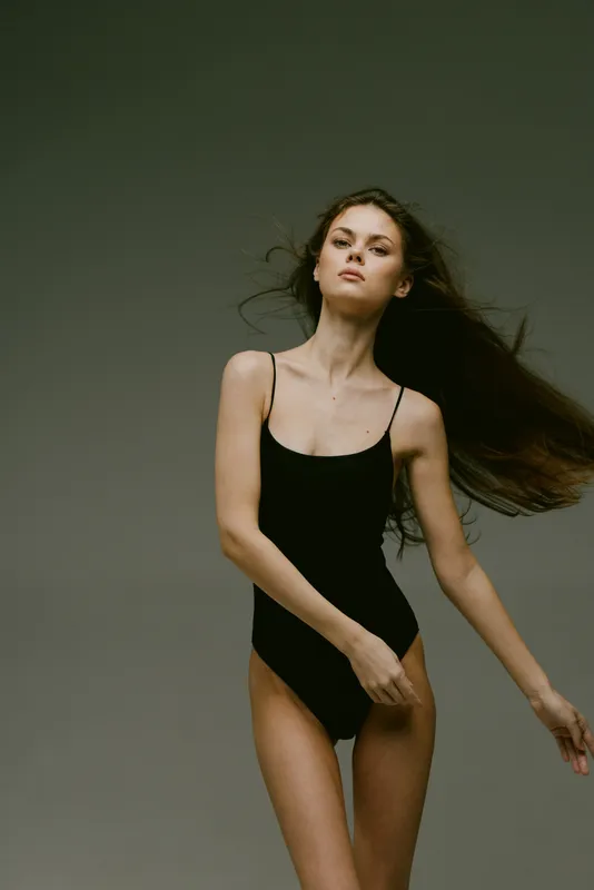 Young woman in black bodysuit posing confidently with flowing hair against gray background