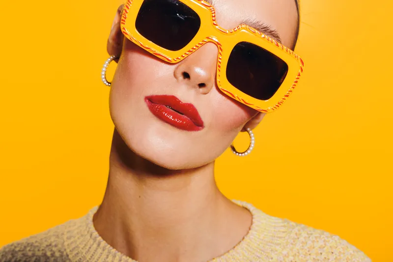 Close-up of a stylish woman wearing large orange sunglasses and red lipstick against a bright yellow background. Concept of fashion, beauty, and summer accessories.