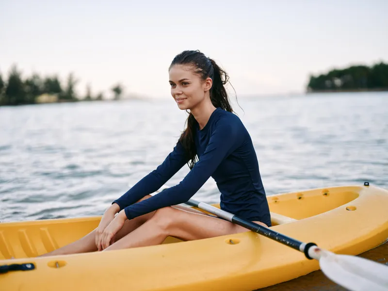 Young woman rowing kayak on calm lake during daytime, wearing sportswear, enjoying outdoor adventure and nature.