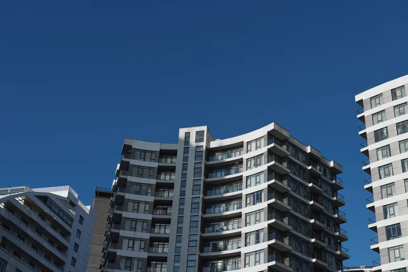 Modern apartment buildings stand tall under a clear blue sky with sunlight highlighting their architectural details in an urban setting.