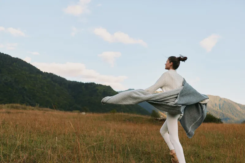 A woman dances in an open field, scarf flowing as wind lifts her dress, capturing a carefree lifestyle and connection to nature. Movement and serenity in outdoor scenery.