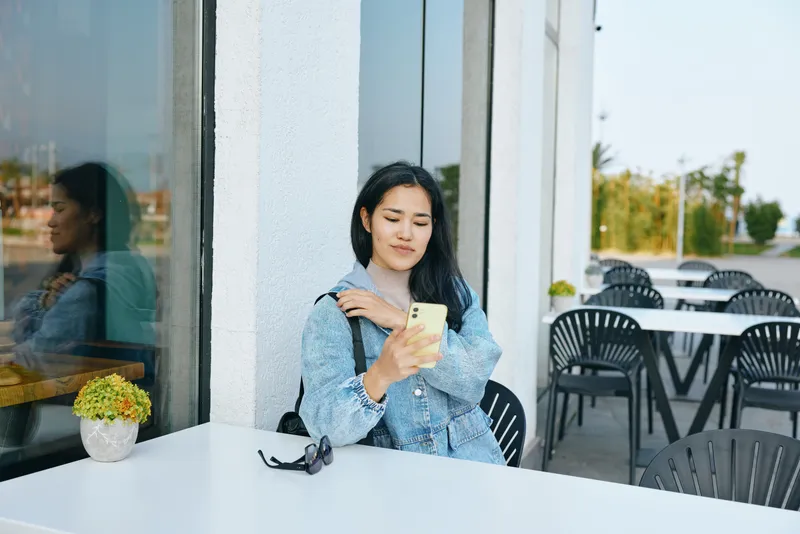 Woman sits at an outdoor cafe, scrolling on her smartphone with a calm casual pose, sunlight on a white table, plant pot, reflective glass, contemporary moment.