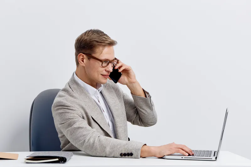 Professional man in formal blazer and glasses sitting at desk with laptop talking on mobile phone isolated on plain white background studio portrait. Business, communication, technology concept.