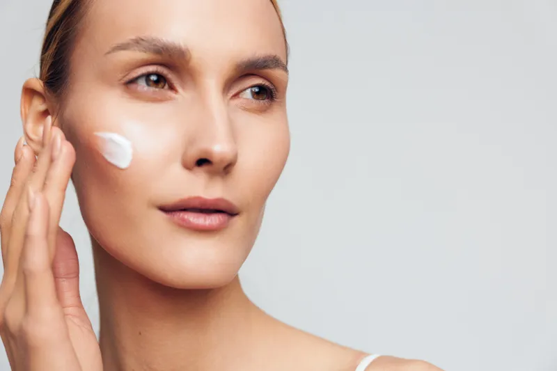 Close up of a woman applying moisturizer on her cheek, clear skin, soft light, skincare routine, calm expression, gentle touch, minimal makeup, isolated on light background, healthy glow