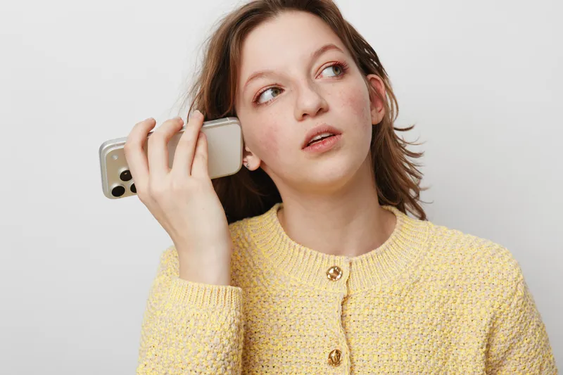 Young woman in yellow sweater holds smartphone near ear, looking away with thoughtful expression. Concept for communication, technology, and contemplation in casual lifestyle.