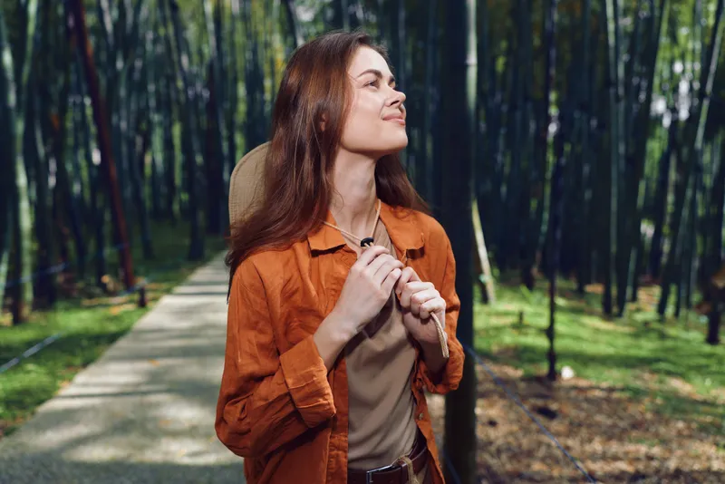 Woman in nature walking on a forest path, smiling and looking up with a hat and casual orange jacket. Outdoor portrait with relaxed travel mood and dappled sunlight.