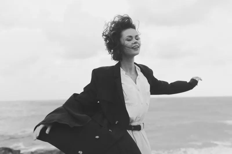 Wind catches a coat and white shirt as a woman dances by the sea, conveying freedom, confidence, outdoor style and dynamic motion.