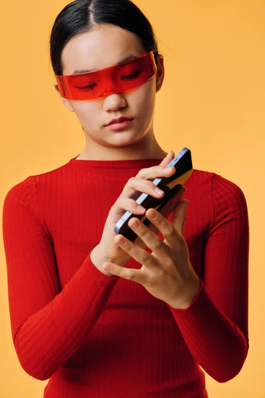 Young woman wearing red top and futuristic red glasses holding black smartphone with focus on screen, isolated on orange background. Female person texting or using mobile device.