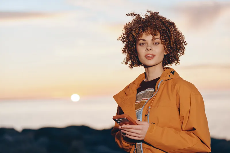 Woman with curly hair in orange jacket holding smartphone at sunset, outdoors near coast, casual lifestyle portrait, warm golden sky light, relaxed mood for travel and connection, by sea