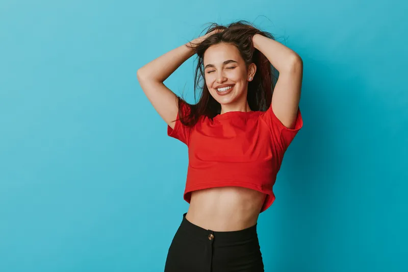 Young woman with closed eyes smiling and hands in hair, wearing red crop top and black pants, posing against blue background for lifestyle and happiness concept.