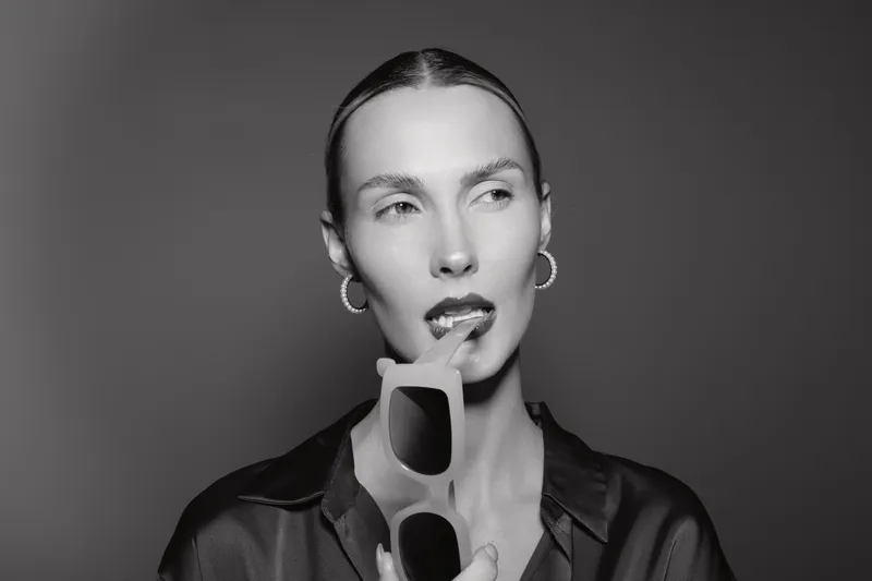 Portrait of a slender caucasian woman with pale skin holding sunglasses near lips, wearing hoop earrings and a dark shirt. Concept of fashion, elegance, and modern style in black and white.