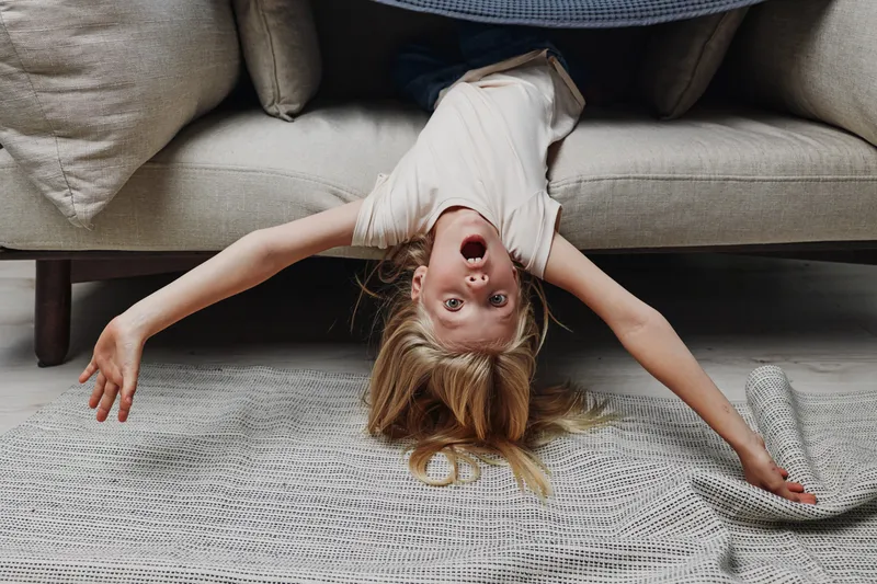 Playful child with blonde hair hangs upside down from a couch, arms outstretched on a knitted rug. Bright indoor scene captures joy, energy, and carefree childhood moments at home.