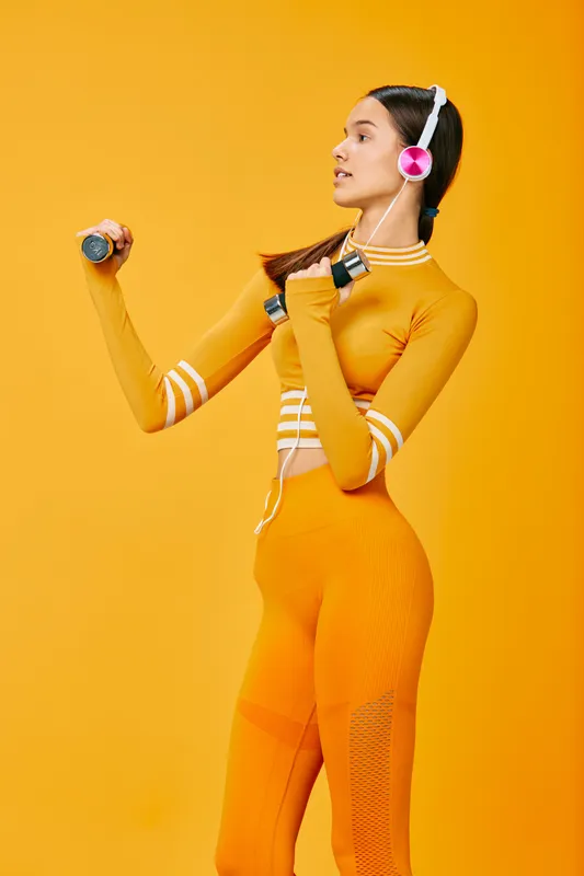 Woman in vibrant orange athletic wear lifting lightweight dumbbells with headphones, engaging in a dynamic fitness routine against a bold orange backdrop.