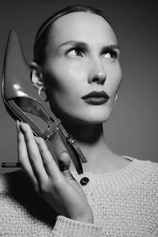 Portrait of a white woman with fair skin holding a high heel shoe near her face, wearing a knitted sweater. Concept for fashion, beauty, footwear, style, and elegance.