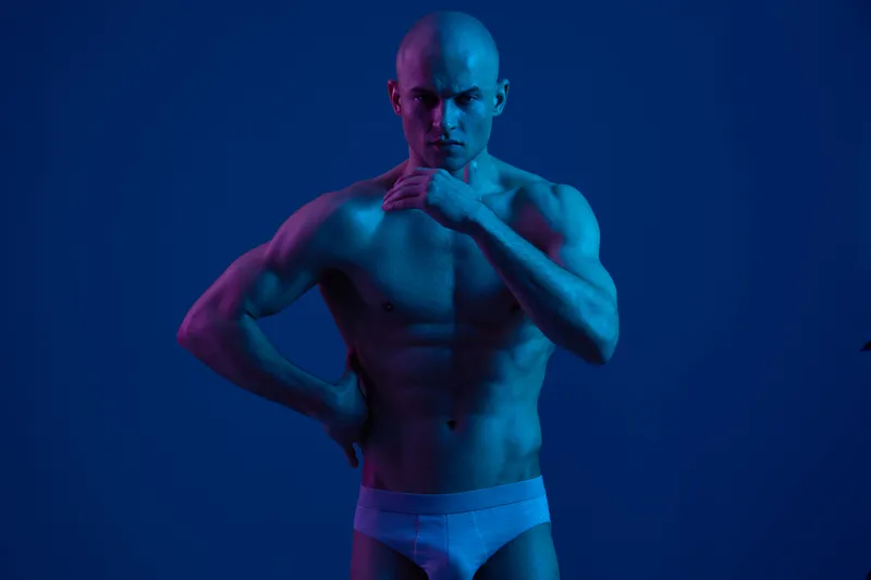 Muscular man posing in dramatic blue lighting, showcasing strength and confidence, emphasizing body art and posture. Ideal for fitness and wellness themes.