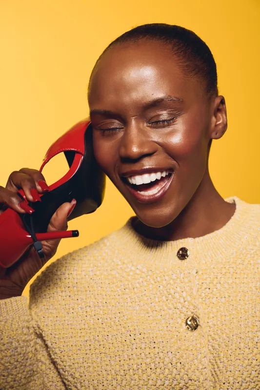Smiling dark skinned African woman with closed eyes holds red high heel shoe near face on yellow background. Concept of fashion, beauty, joy, and elegant accessories in studio.