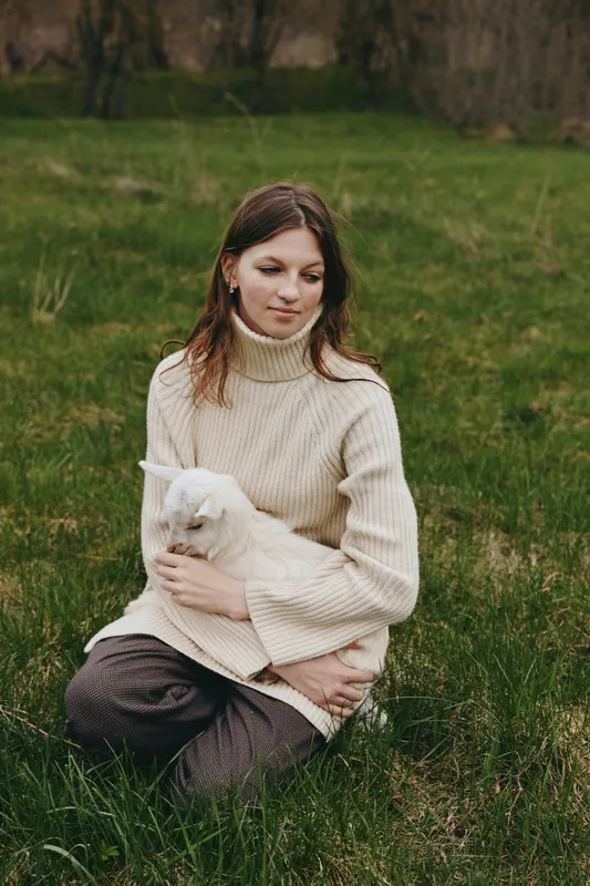 Calm woman in a cream sweater sits on grass, gently cradling a white goat kid in a rural field, lifestyle scene about companionship with animals, natural light creating a warm, peaceful mood