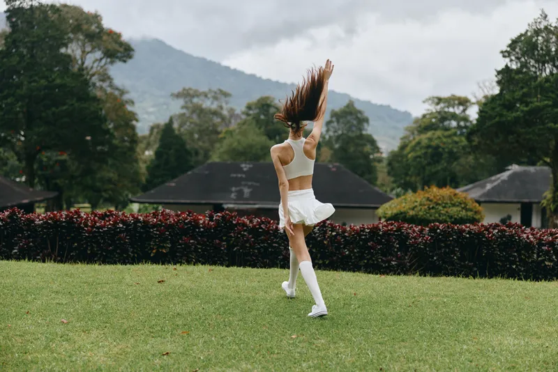 Young woman joyfully dancing in a green park, wearing a white athletic outfit and knee high socks, surrounded by lush nature and mountains in the background