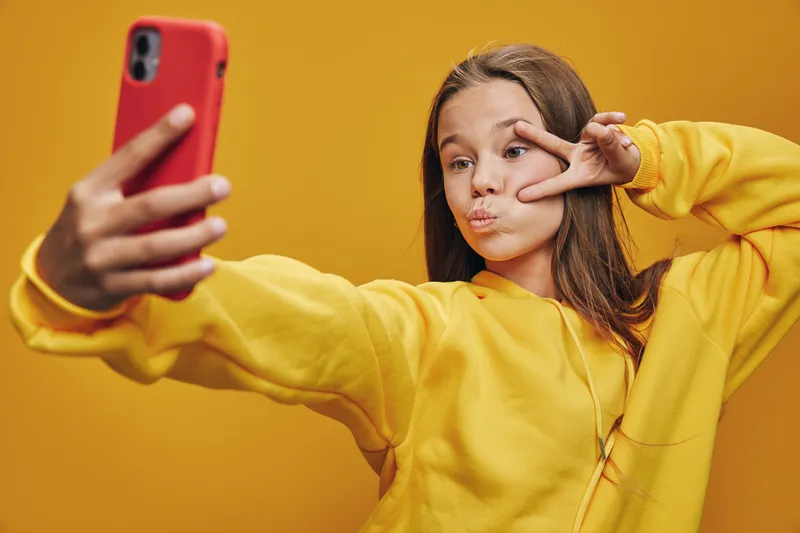 Teen girl in yellow sweater taking selfie with smartphone making peace sign with fingers on orange background. Casual portrait of smiling happy young woman indoors.