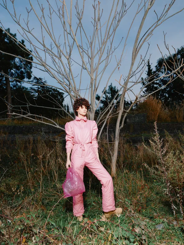 Fashion model in an outdoor setting wearing a pink jumpsuit, standing by a bare tree with moody light, showcasing chic streetstyle and contemporary clothing in a natural scene.