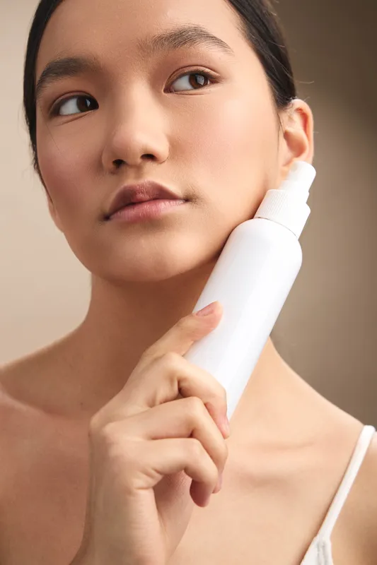 asian woman applies lotion from a white bottle in a softly lit space, showcasing a skincare routine with a calm, confident expression and a clean, modern atmosphere.