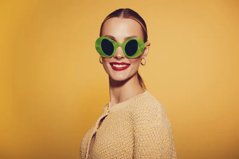 Smiling woman with green round sunglasses and red lipstick poses against yellow background. She wears a beige knitted sweater and gold hoop earrings. Concept of fashion and style.