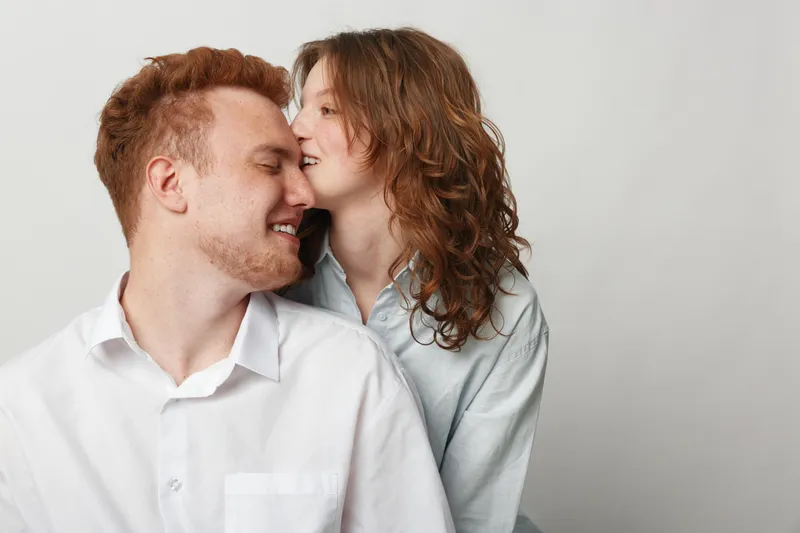 Happy young couple in casual shirts share intimate moment, woman whispering to man with closed eyes. Concept of love, affection, trust, and close relationship in studio setting.