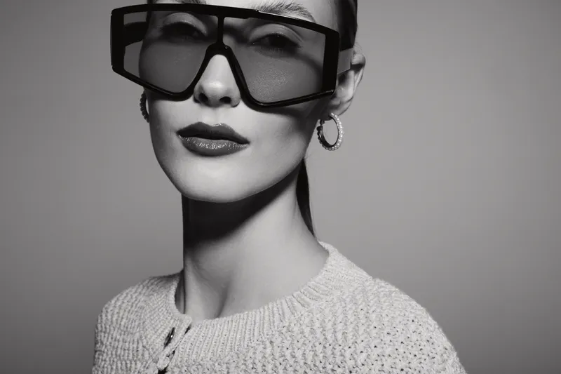 Elegant Caucasian woman with light skin wearing oversized square sunglasses and hoop earrings poses in a knitted sweater on gray background. Fashion and style concept.