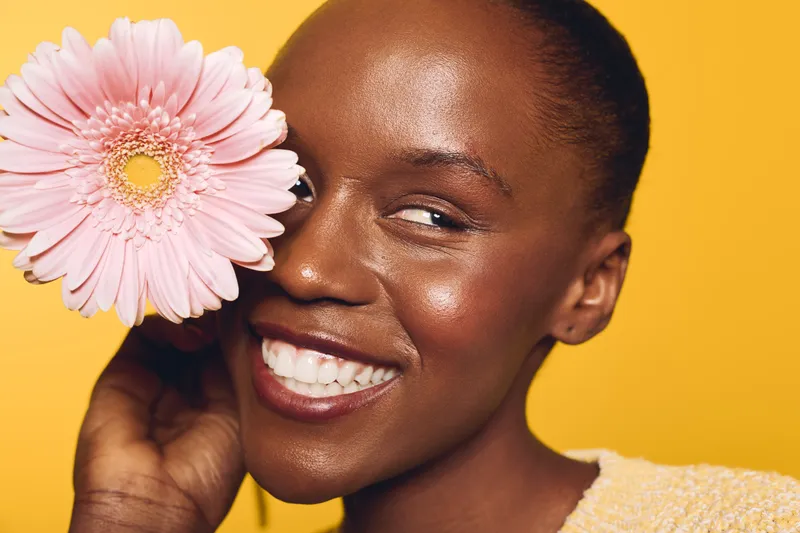 Smiling dark skinned African woman holds pink flower near face on yellow background. Close-up portrait of happy female model with natural skin glow and positive expression. Concept of beauty.