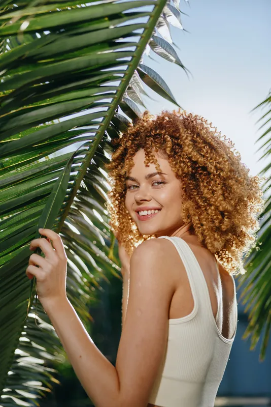 smiling woman with curly hair wearing white top among green palm leaves outdoors, natural light highlighting warm skin tones, summer vibe and fresh atmosphere
