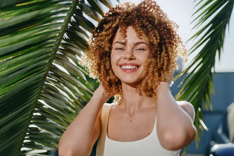 Smiling woman with curly hair enjoys sunny tropical outdoor setting wearing white tank top against palm leaves, radiating happiness and natural beauty.