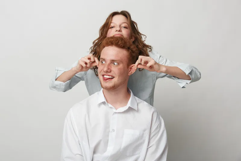 Playful young woman pulls ears of smiling man in white shirt against plain background. Concept of friendship, fun, humor, and casual lifestyle with happy emotions and light mood.