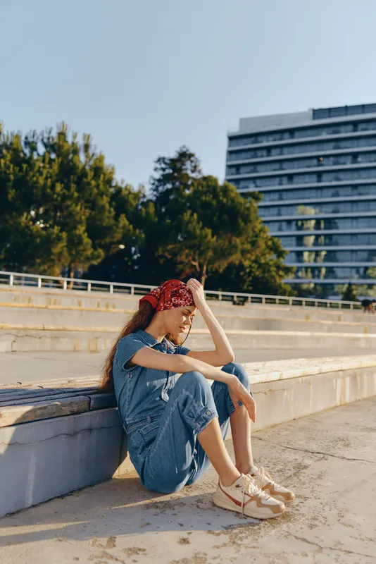 Lifestyle woman in modern boho-western denim street style with a red bandana, sitting outdoors by concrete steps under bright natural light, film color effect.