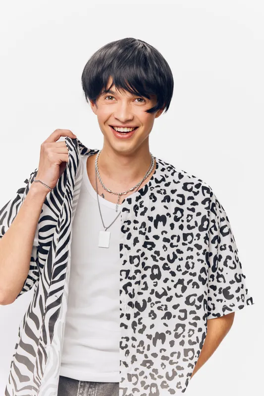 smiling man in patterned shirt holding jacket over shoulder with casual hairstyle and layered necklaces, isolated on white background, youthful and fashionable style