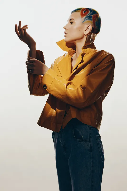 Woman with colorful patterned hair wearing a mustard yellow jacket and jeans poses thoughtfully against a soft neutral background with artistic lighting.