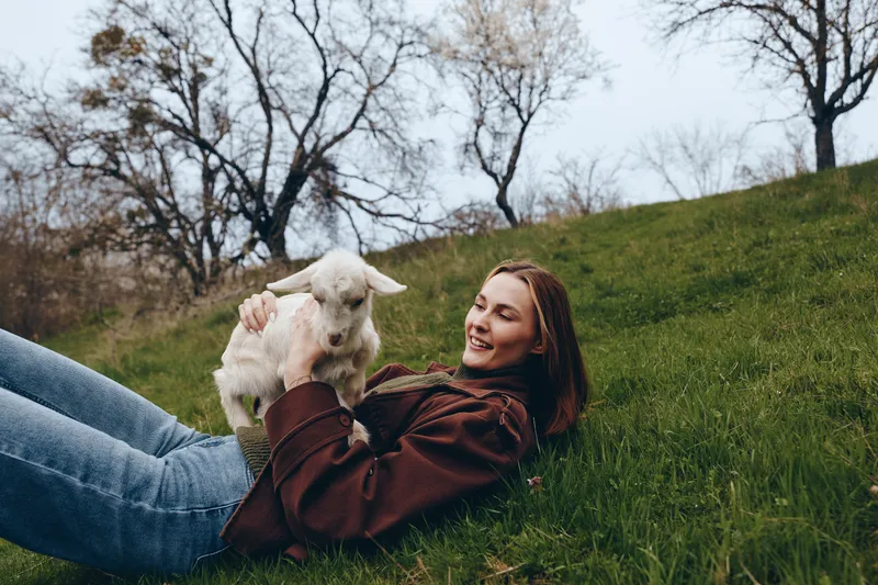 Relaxed woman lying on grassy hillside with a playful goat kid nearby, outdoors scene, casual jacket, gentle breeze, natural colors, intimate lifestyle moment in nature