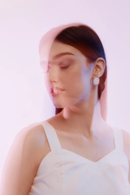Young woman in white dress poses with eyes closed and blurred double exposure effect. Soft pink and purple lighting highlights her beauty and fashion style. Studio portrait.
