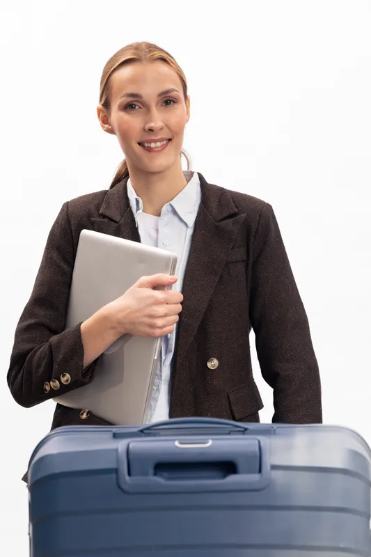 Smiling young businesswoman holding laptop stands behind blue suitcase isolated on white background. Concept for travel, business trip, and professional woman on the move.