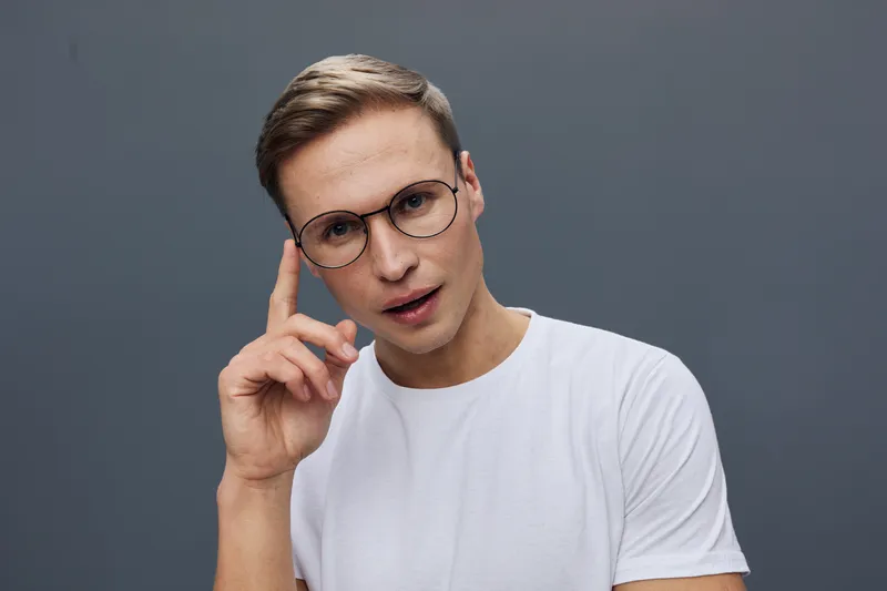 Thoughtful young man with glasses wears white t-shirt look camera hold in hand isolated on plain solid gray background studio portrait. People lifestyle concept