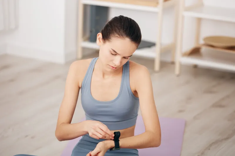 Young woman in sportswear adjusts a smartwatch while seated on a purple yoga mat at home, preparing for fitness training; calm indoor minimalist interior and focused posture.