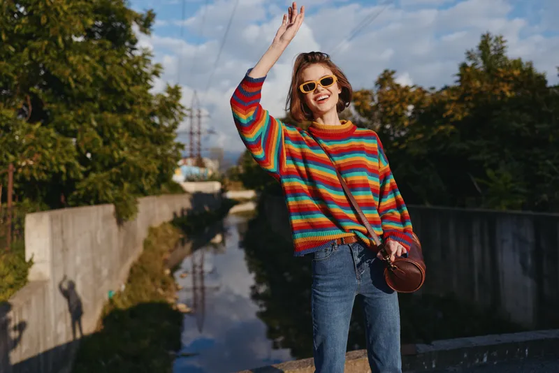 Woman in striped sweater and sunglasses waving and smiling by urban canal, wearing jeans and crossbody bag, casual outfit, young adult traveler enjoying sunny outdoor lifestyle moment.