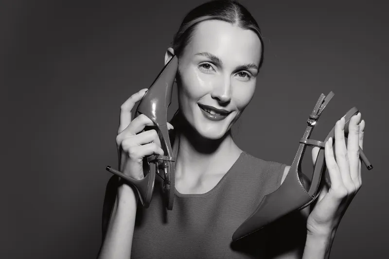 A smiling Caucasian woman with fair skin holds red high heels near her face in a studio with a dark background. Concept for fashion, style, and footwear.
