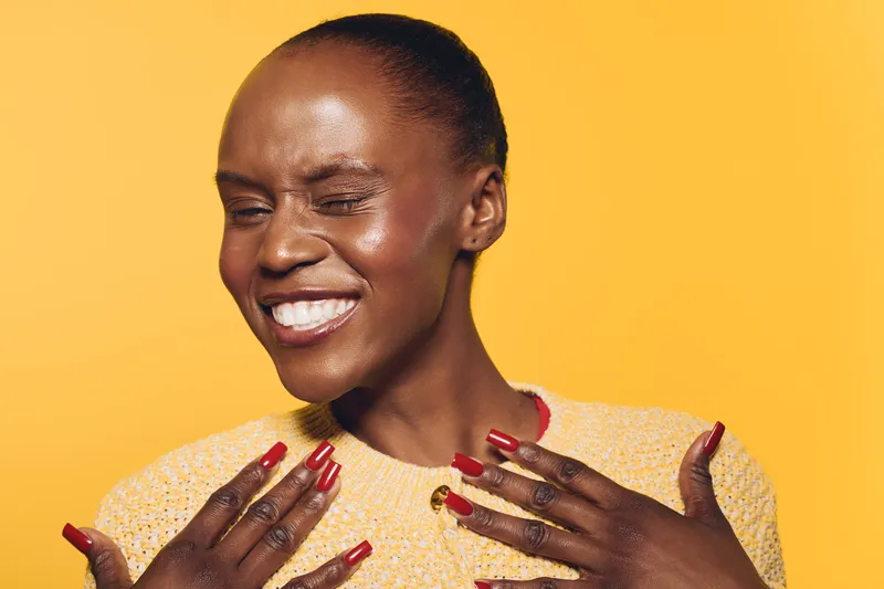 Joyful dark skinned African woman with short hair smiles with closed eyes on yellow background. Close-up portrait with red nails and happy expression. Concept of beauty and positive emotions.