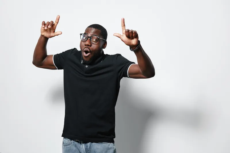 African American man with dark skin wearing glasses and black polo shirt making excited gesture with hands and surprised facial expression on white background.